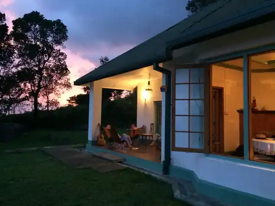 The Bungalow's lamp-lit veranda and open windows at dusk under a pink sky