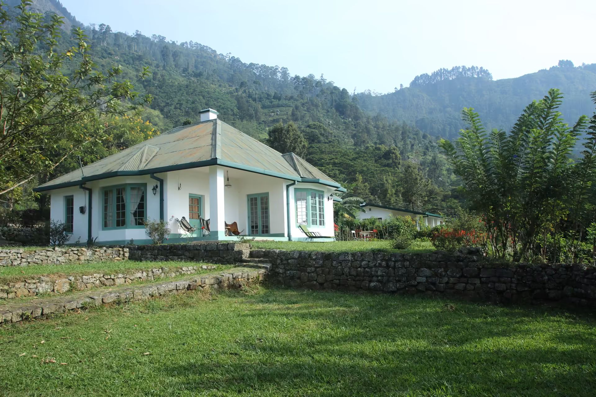 The Bungalow, a white planter's house with a green roof and bay windows, above its terraced lawn and stone walls, with forested hills behind
