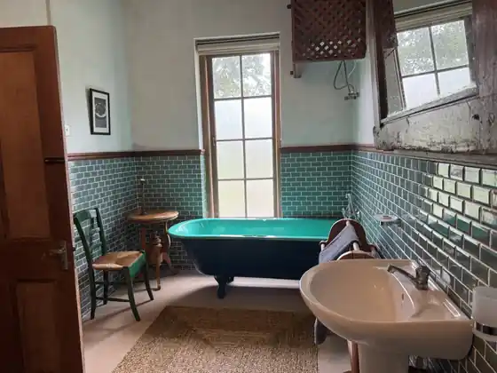 A Bungalow bathroom with a green roll-top bath under a tall window, small green tiles and a pedestal basin